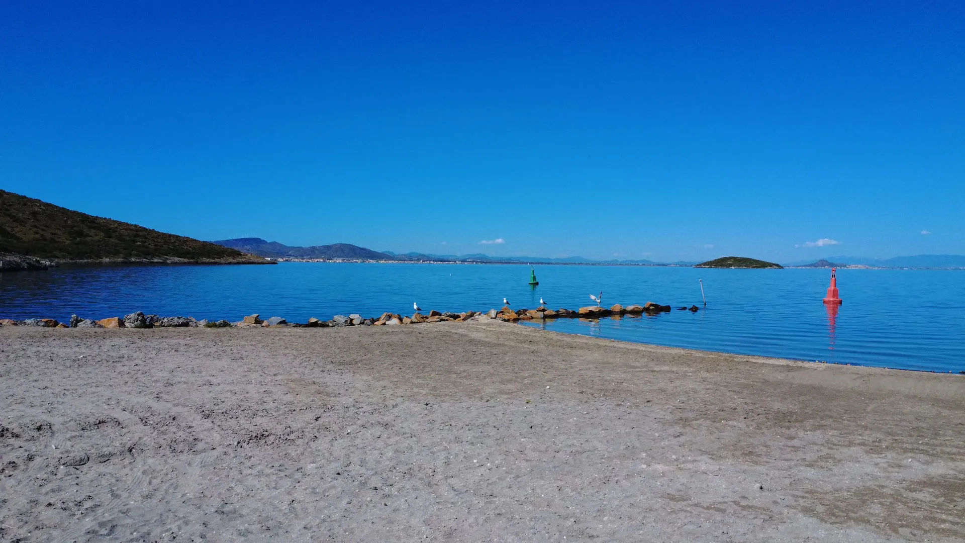 Sandy beach with a rock jetty extending into calm blue water, green and red navigational buoys, distant hills, and clear blue sky.