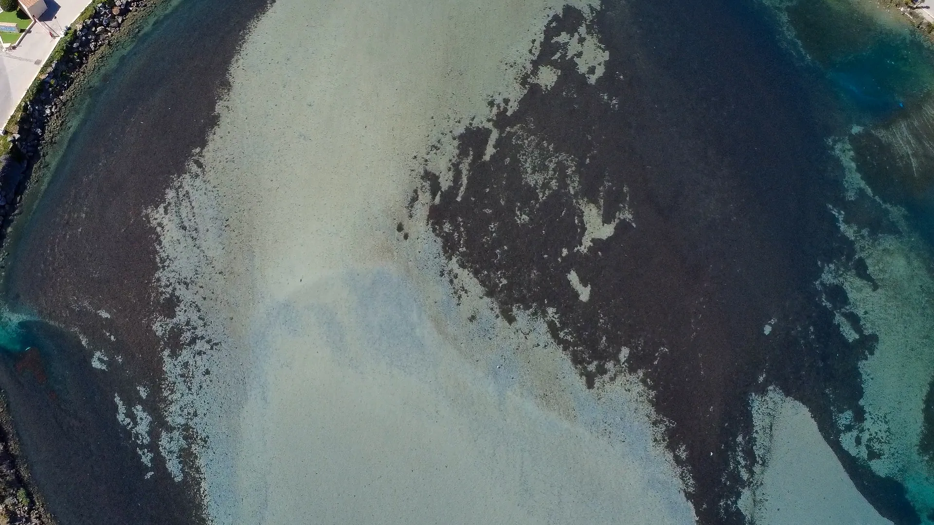 Aerial view of shallow coastal waters displaying varying shades of blue and brown, with rocky edges and sandy patches.