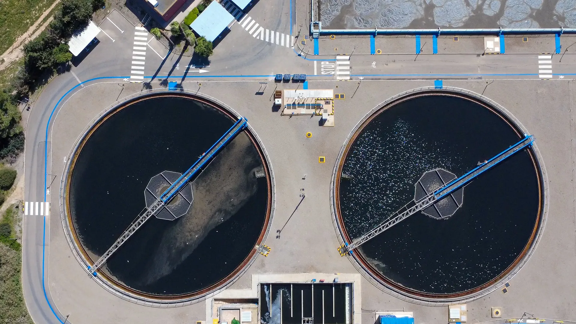 Aerial view of two large, circular water treatment tanks, with a walkway and machinery above, surrounded by roads and buildings.